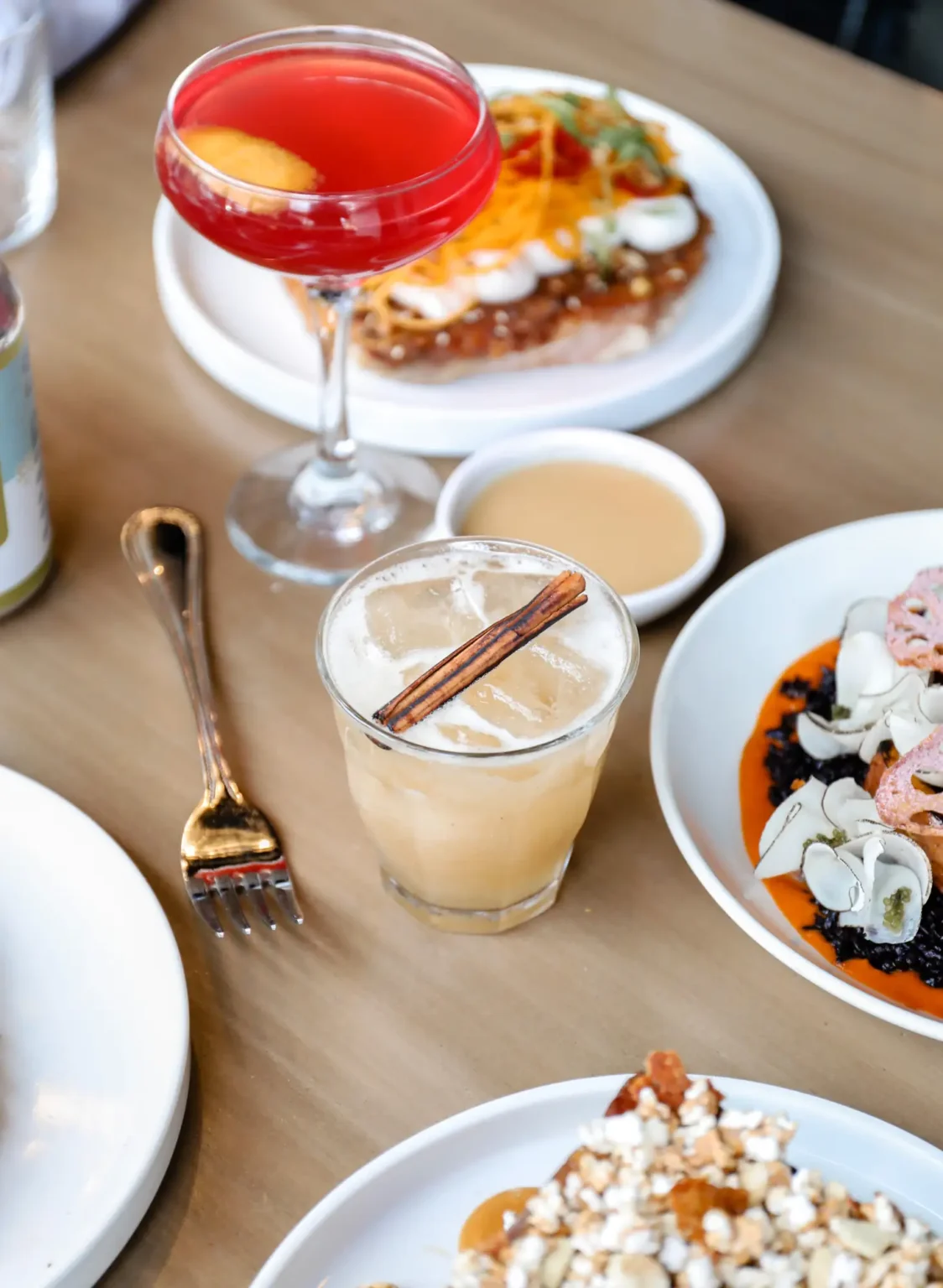 A brunch table seen from above with a pink coupe cocktail, an iced drink with a cinnamon stick and plates of toast