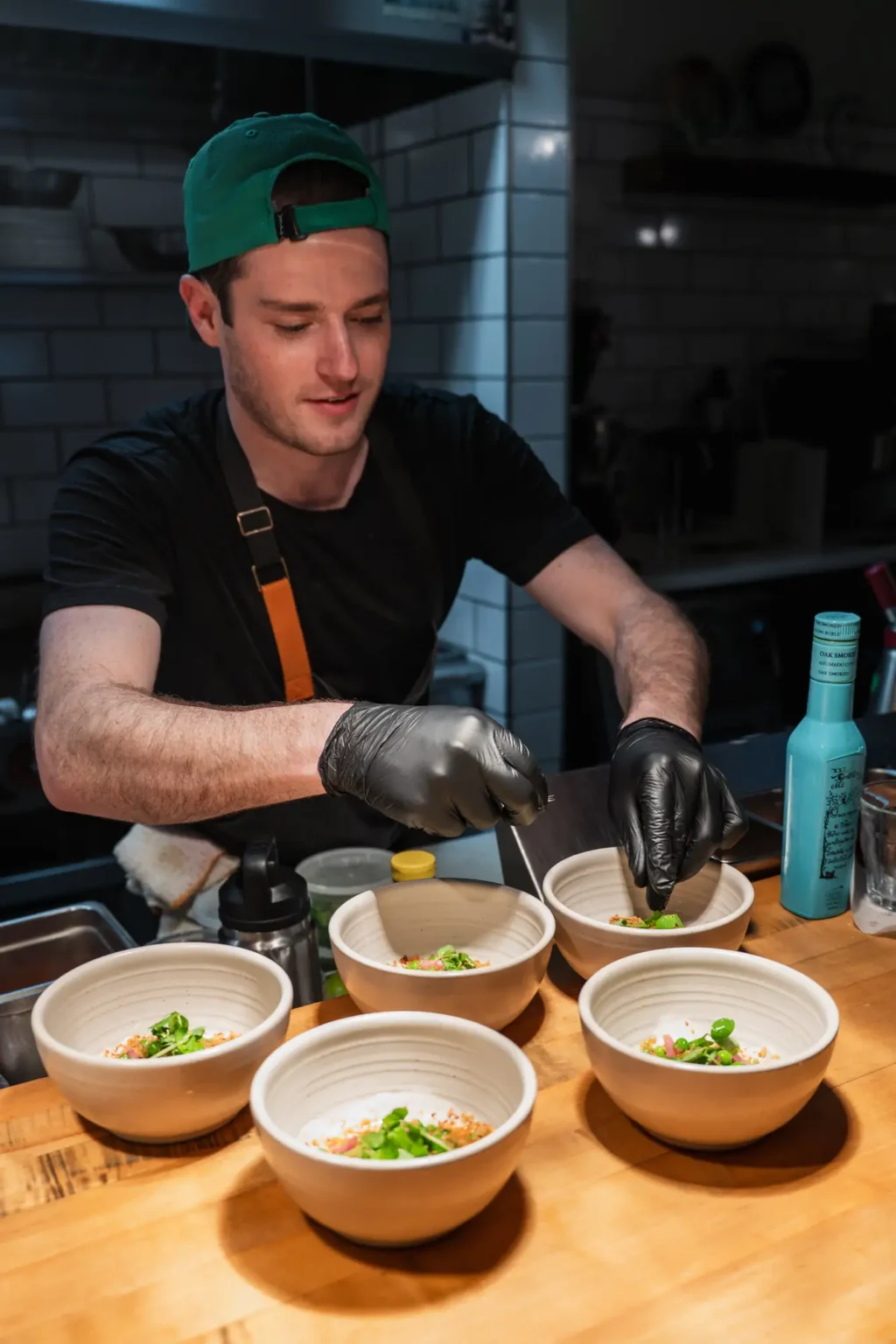 A chef in a green cap garnishing a row of small bowls at the kitchen pass