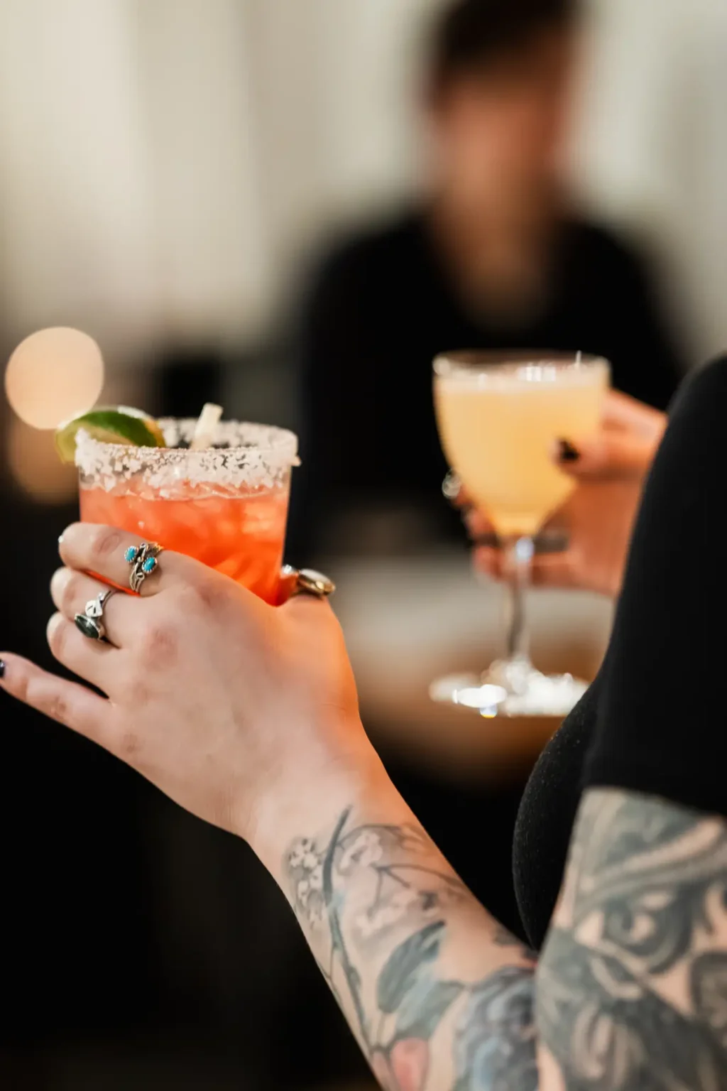 Two guests raising cocktails, a salt-rimmed red drink and a pale coupe
