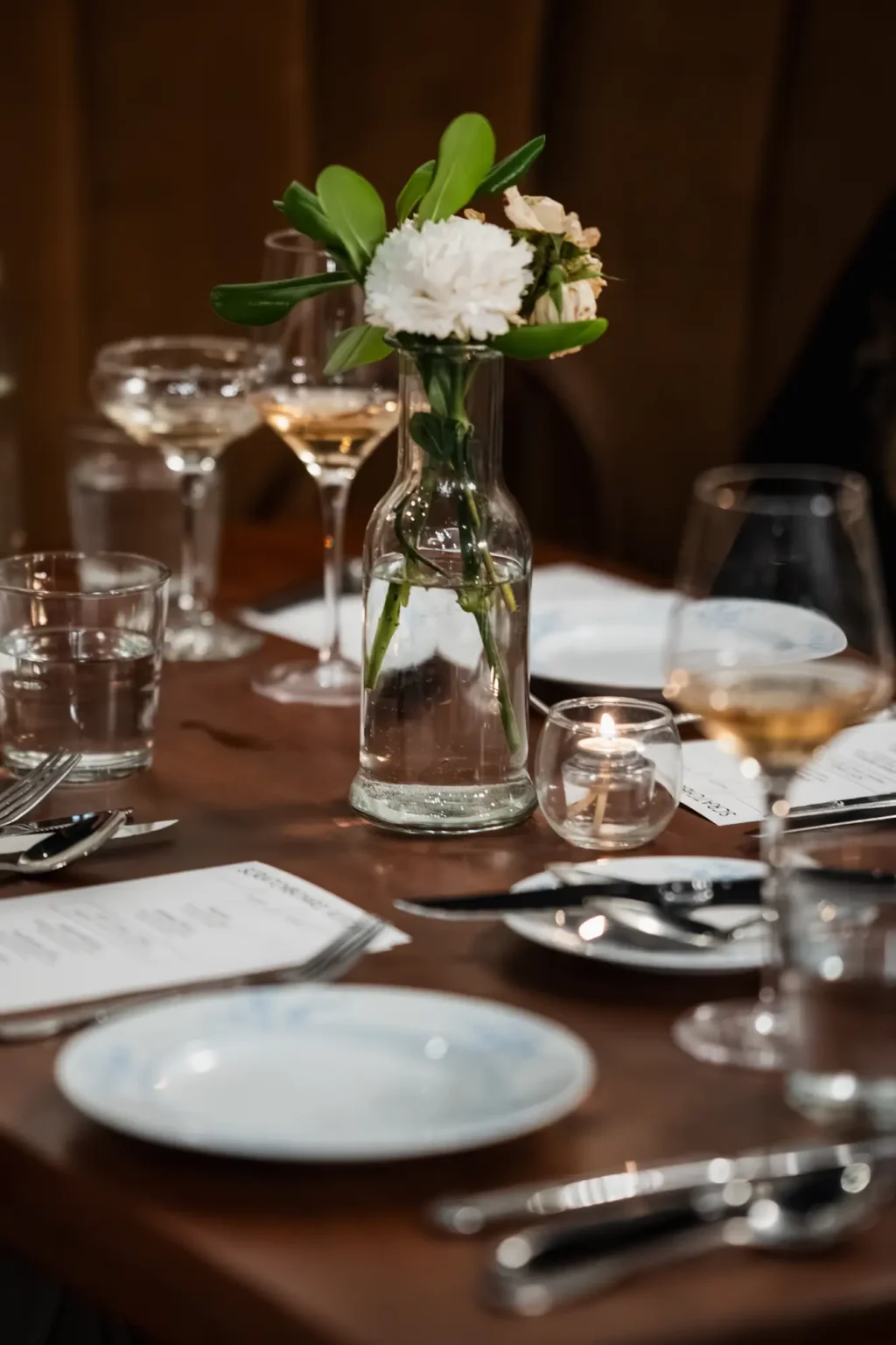 A candlelit table set for dinner with a white carnation in a bud vase and glasses of white wine