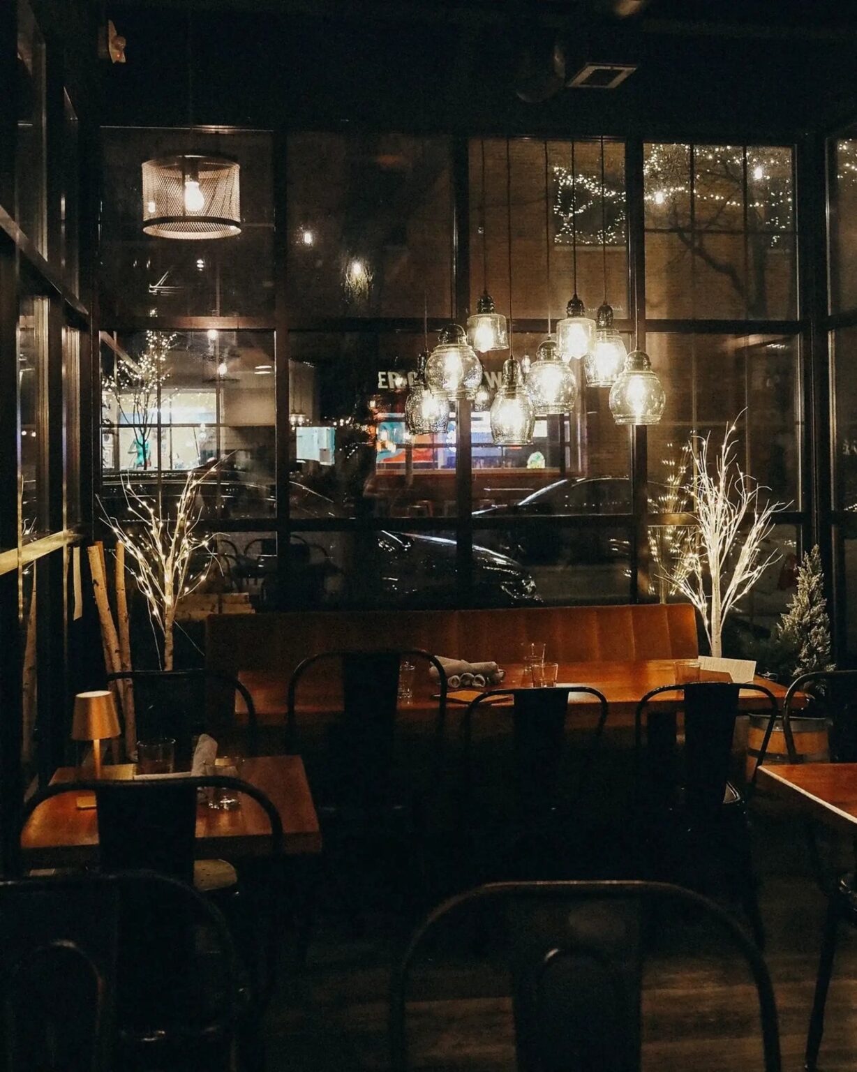 The Scratchboard Kitchen dining room at night, with glowing pendant lights, wooden tables and tall windows onto the street