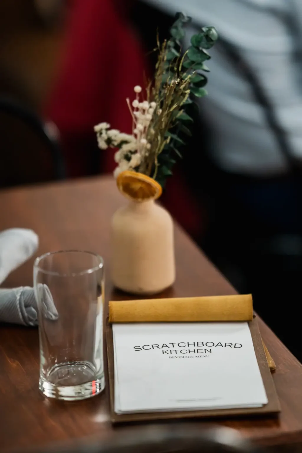A Scratchboard Kitchen beverage menu on a table beside a small bud vase