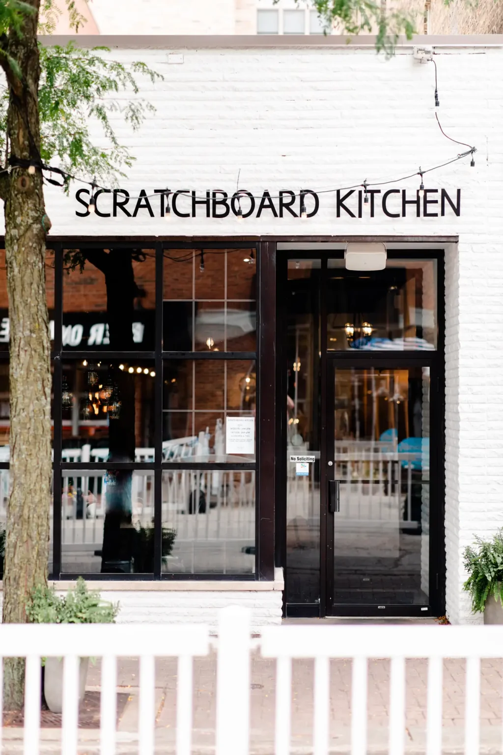 The front entrance of Scratchboard Kitchen, a white-brick building with the restaurant's name above tall black-framed windows