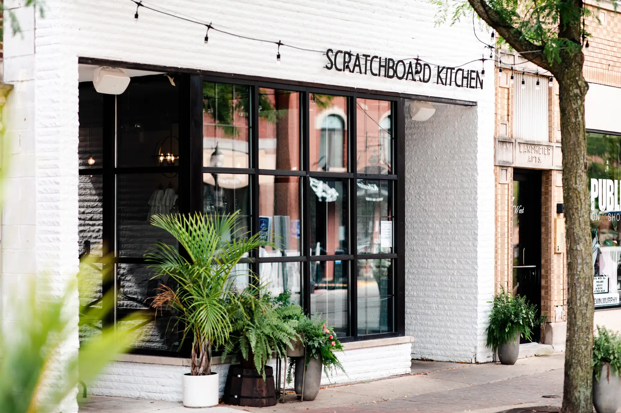 The white-brick storefront of Scratchboard Kitchen on Campbell Street, with tall black-framed windows and potted palms by the door