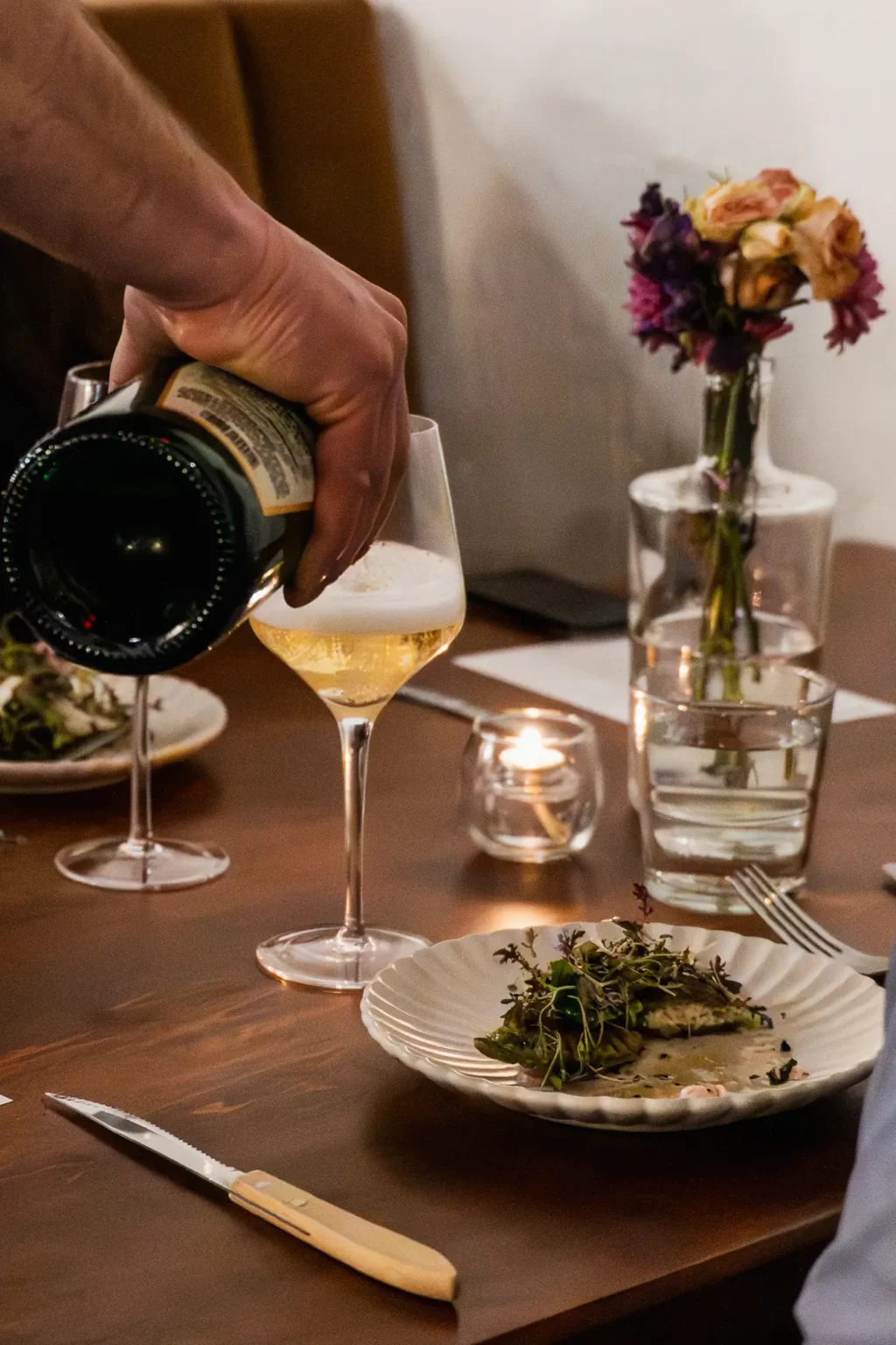 A server pouring sparkling wine beside a plated dish and a small vase of flowers